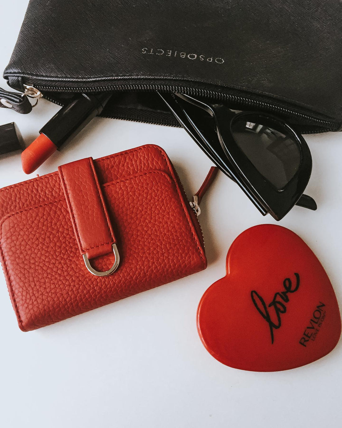 Red leather wallet with a strap closure beside sunglasses, lipstick and a heart shaped mirror