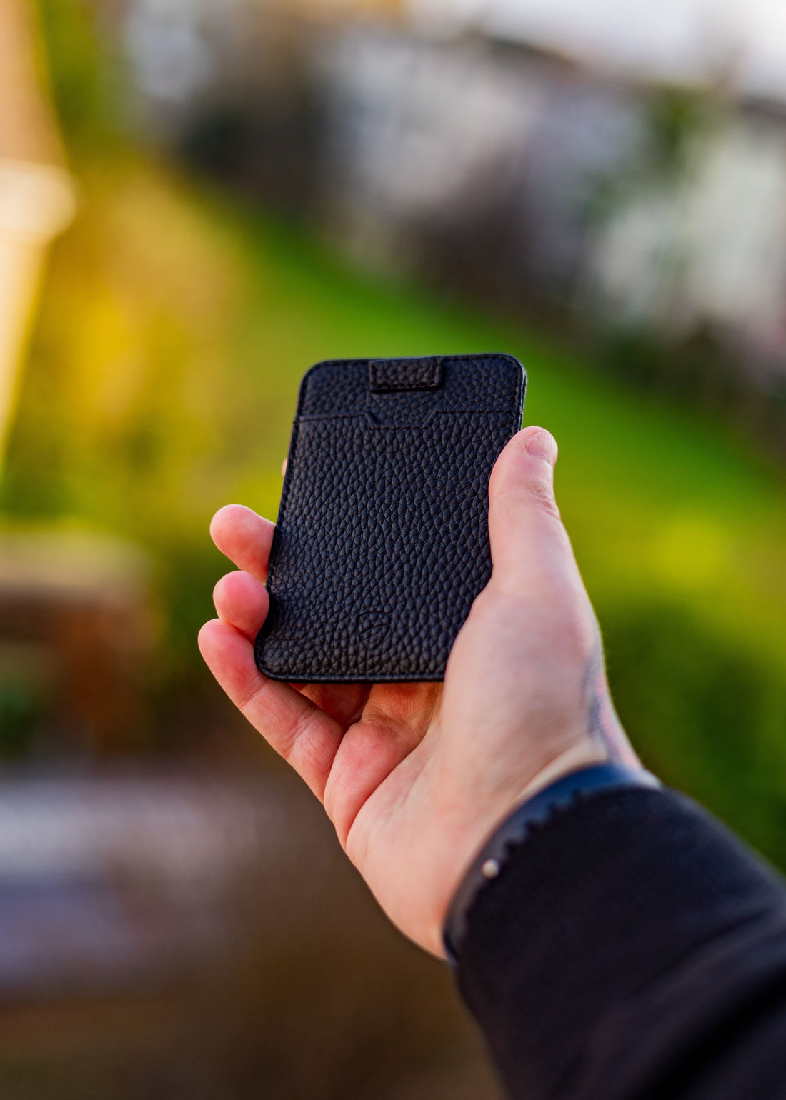 A hand holds the black leather CHELSEA wallet outdoors against a blurred green background