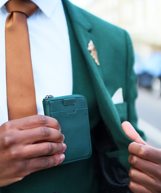 Man in a green blazer holding a green leather NOTTING HILL zip-around wallet with a tan tie visible