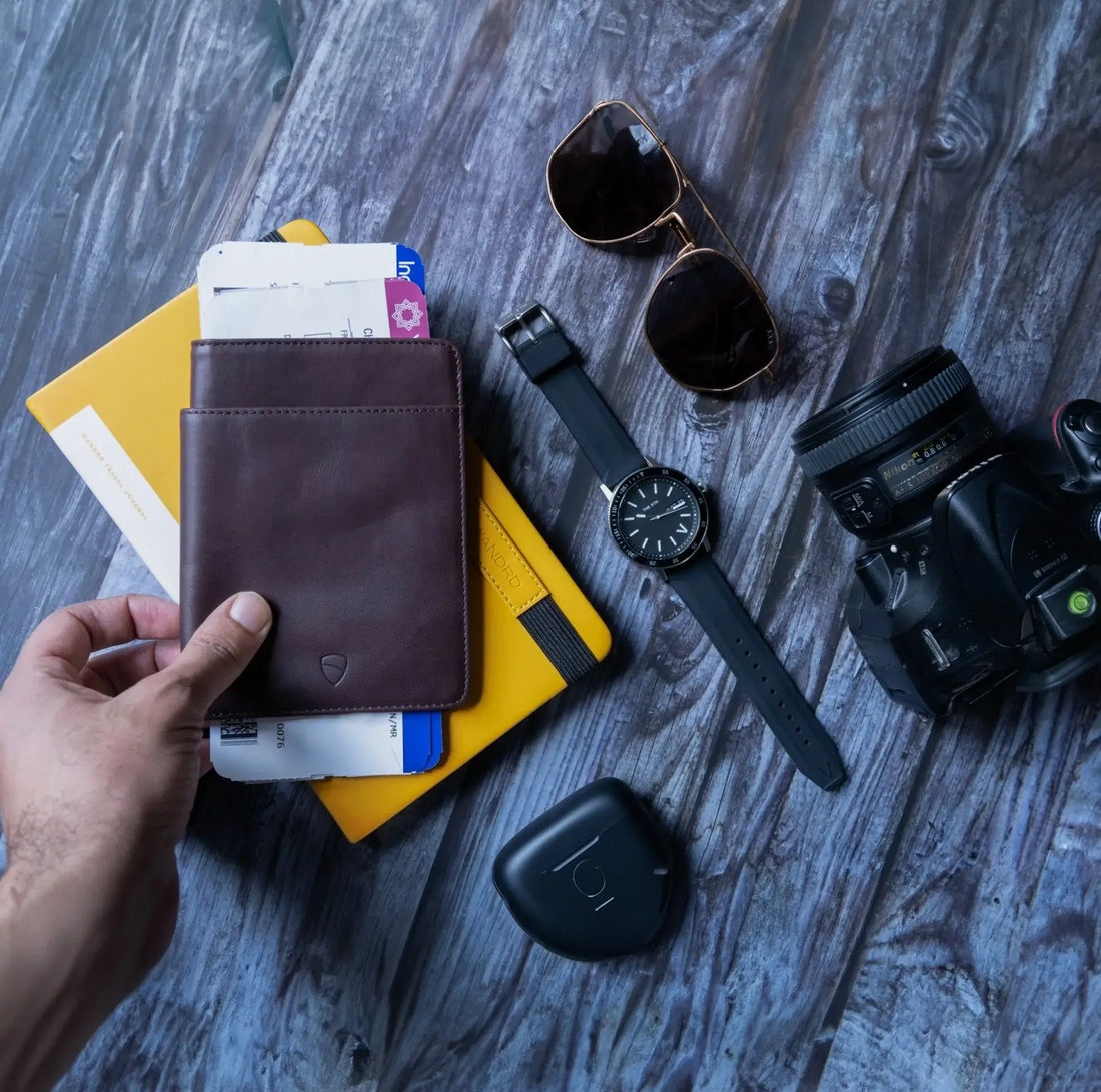 A hand holds a brown KENSINGTON passport wallet with cards on a yellow notebook beside sunglasses, a watch, a camera and a tracker tag