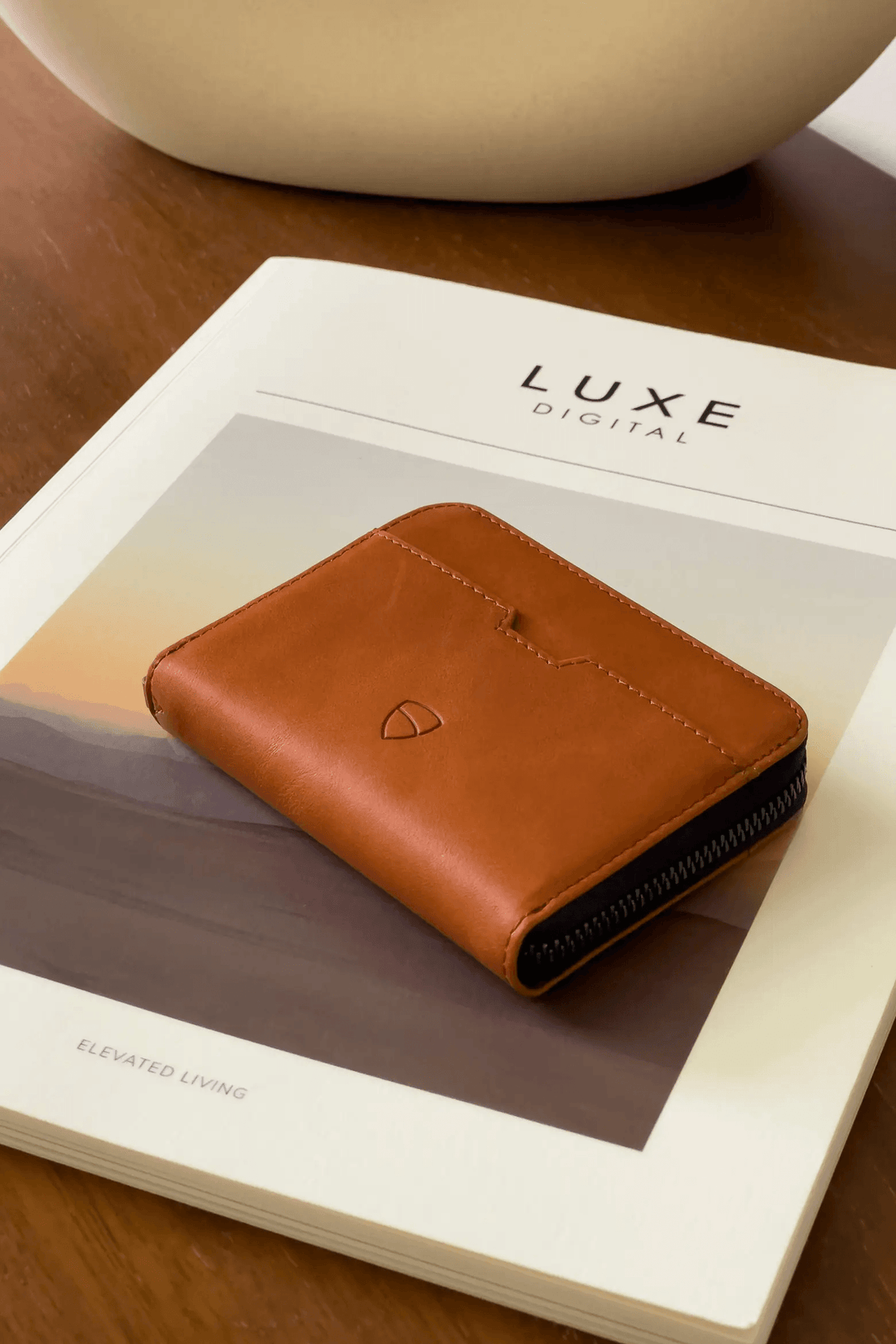 A tan leather zip-around wallet resting on a magazine cover beside a ceramic vase on a wooden table