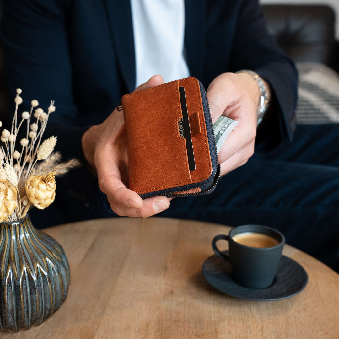 A man in a suit holds a tan leather zip-around wallet with card slots and a folded banknote beside a coffee cup