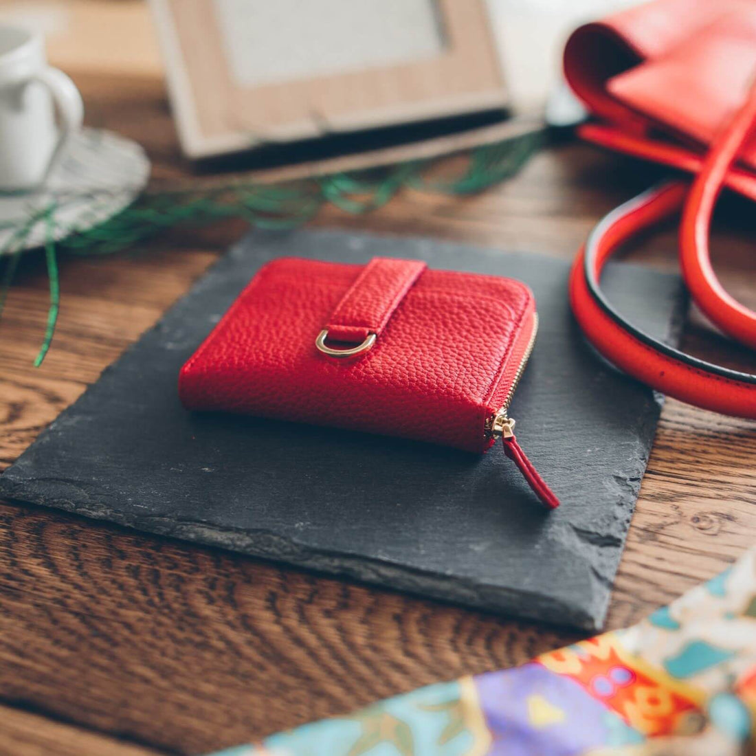 A small red pebbled leather zip wallet with a tab strap sits on a dark slate tile