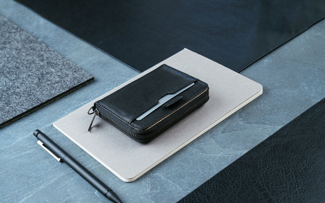 A black leather MAYFAIR zip wallet with a card peeking out, resting on a notebook beside a pen