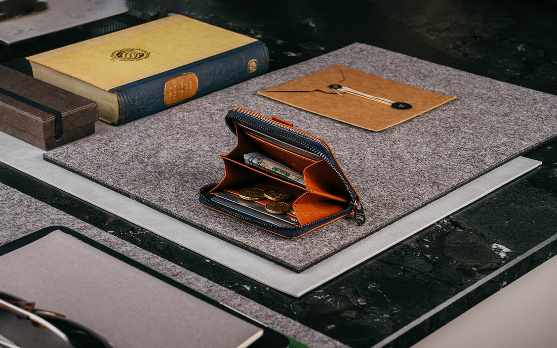 An open cognac and navy MAYFAIR zip wallet with coins and a banknote inside, beside a book and envelope