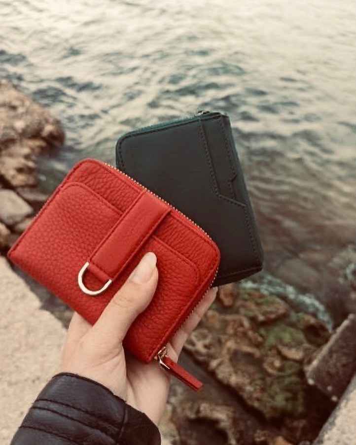 A hand holds a red NOTTING HILL wallet with its Smart Strap over a black leather zip wallet above water