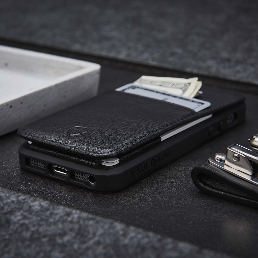 A black leather phone case holds banknotes and a card above its charging port and headphone jack, beside a nail clipper on a dark surface