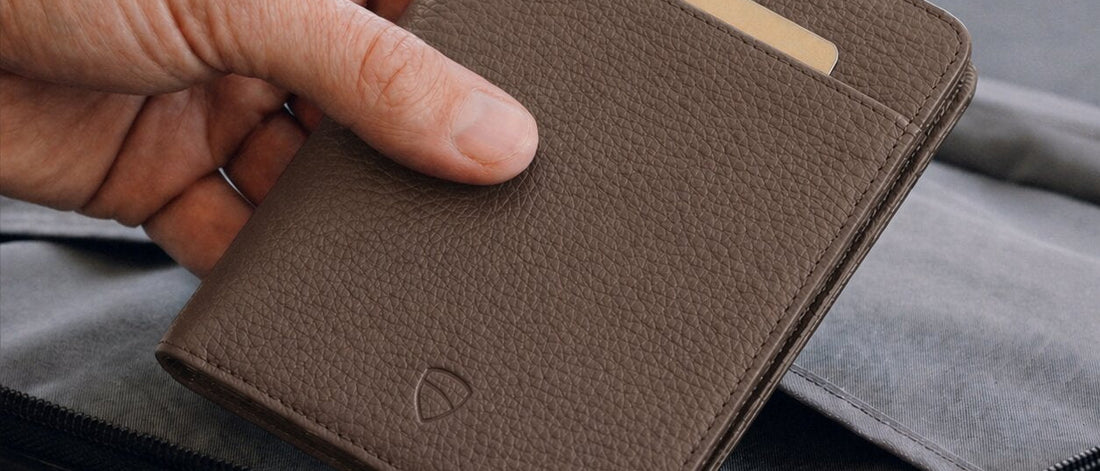 A hand holds a brown KENSINGTON passport holder with a card tucked into its slot above a suitcase