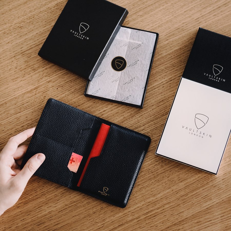 A hand opens a black leather KENSINGTON passport wallet with red interior beside its packaging boxes