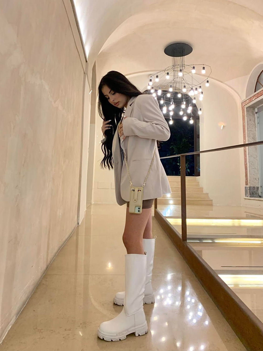 Woman in a beige blazer carries a cream phone case on a gold chain through a marble hallway