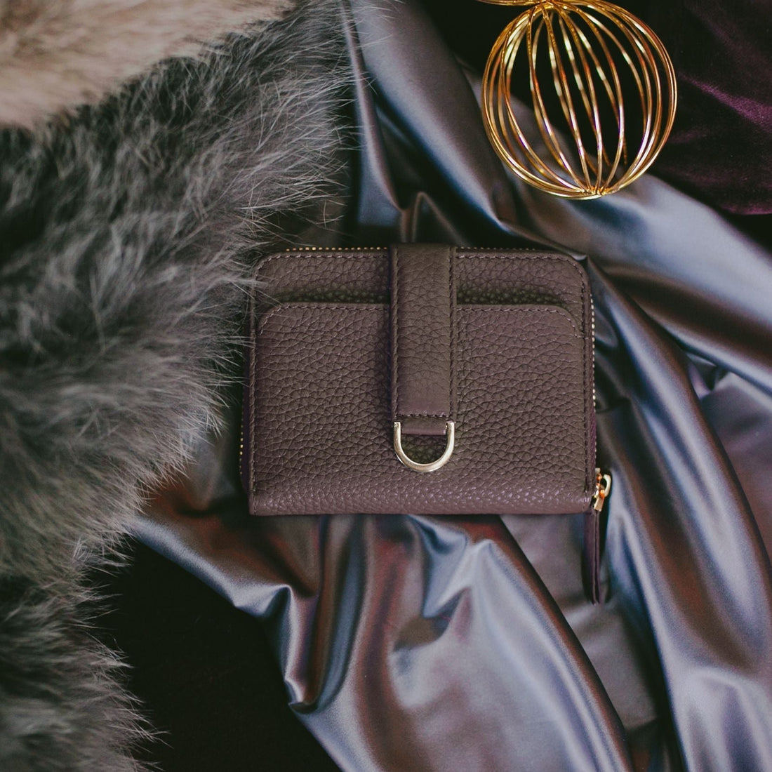 A brown leather zip-around wallet with a gold strap rests on silky fabric beside fur and a gold wire sphere