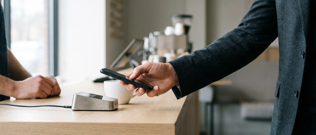 A customer pays by holding a smartphone to a contactless card terminal on an oak café counter beside a white cup