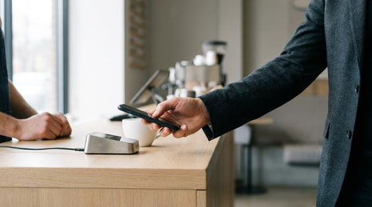 A customer pays by holding a smartphone to a contactless card terminal on an oak café counter beside a white cup