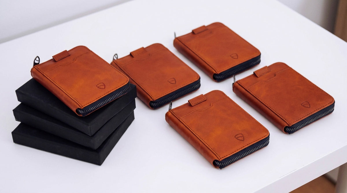 5 hellbraune Reißverschluss-Geldbörsen aus Leder mit Zuglaschen liegen neben einem Stapel von 3 schwarzen Geschenkboxen auf einem weißen Tisch